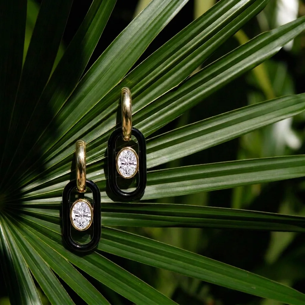 Oval hoop earrings with black enamel links resting on a fanned palm leaf in sunlight